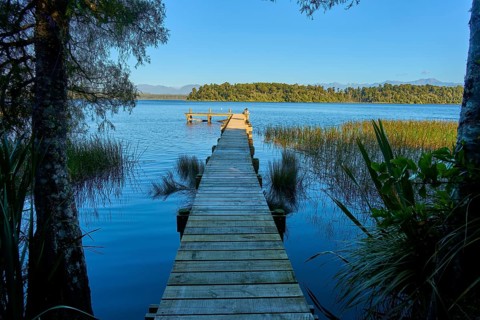 A wooden footbridge leads from a wooded shore into a calm lake, Lake Mahinapua, Ruatapu, South