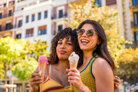 Two smiling girls are eating ice cream cones, hugging each other in a city center during a sunny