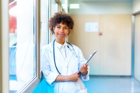 Female doctor standing confidently in a bright hospital corridor, holding a medical chart and