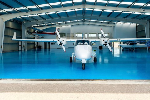 White twin engine airplane parked inside a spacious hangar, featuring a bright blue floor and