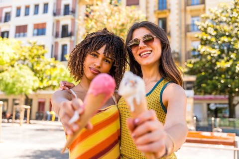 Two friends enjoying a sunny day in the city, happily holding and sharing colorful ice cream cones