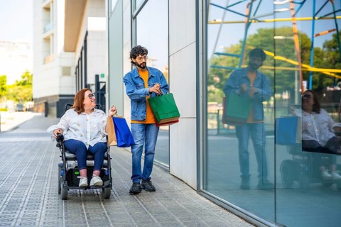 Couple walking on city sidewalk after shopping, woman using a wheelchair and carrying colorful bags