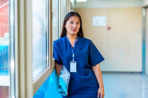 Confident young medic holding a clipboard and wearing blue scrubs is standing next to a window in a