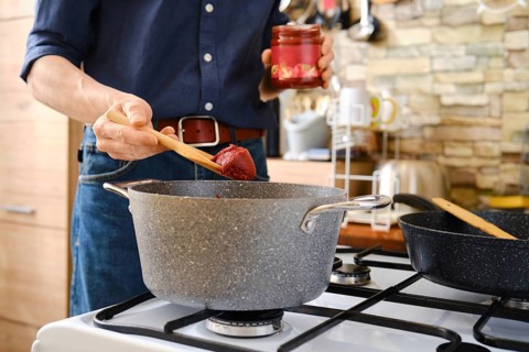 A person prepares a homemade meal adding tomato paste to a pot on the stovetop in a bright, modern