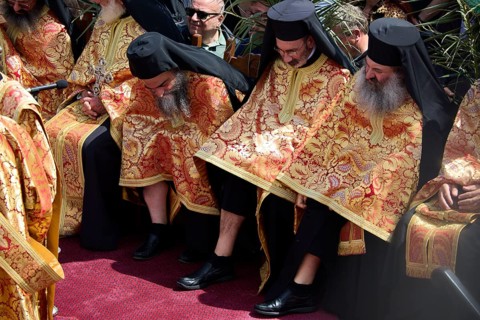 Orthodox priests in elaborate golden robes sit in the middle of a crowd during a ceremony, Orthodox
