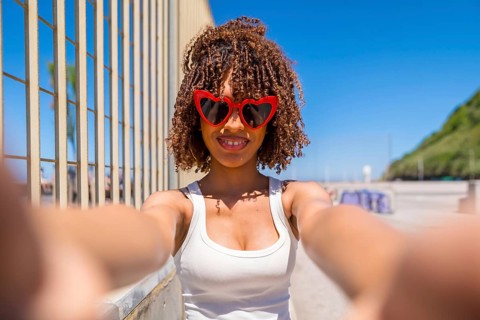 Smiling young woman with curly hair taking a selfie with heart shaped sunglasses on a sunny beach