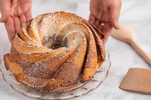 Hands present a bundt cake dusted with icing sugar on a glass plate