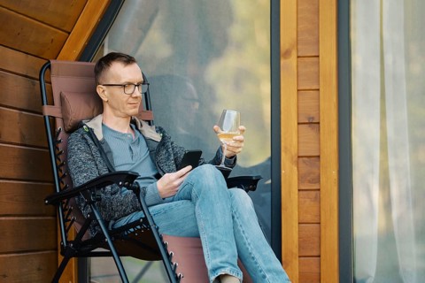 Middle-aged man looking at smartphone sitting on terrace of a house with a glass of wine