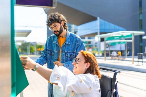 Woman in a wheelchair validating her ticket at a modern tram stop, with a friend providing