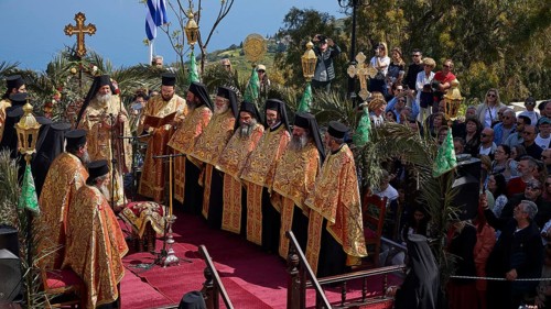 Priests in golden robes lead a religious ceremony on a decorated stage in front of a Greek flag and