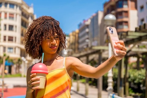 Young woman taking a selfie with her smartphone while holding a milkshake, enjoying a sunny day in