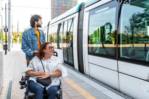 Caregiver pushing a smiling woman in a wheelchair at a modern tram station, both eagerly waiting to
