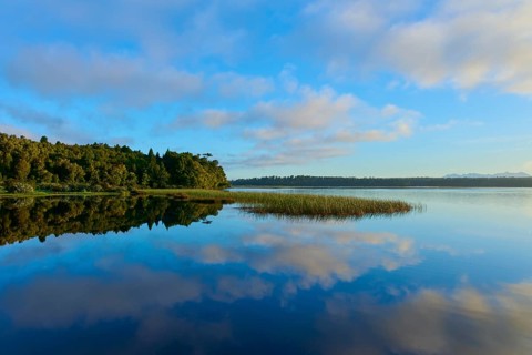 Blue sky and clouds reflected in a calm lake with a forested shore, Lake Mahinapua, Ruatapu, South