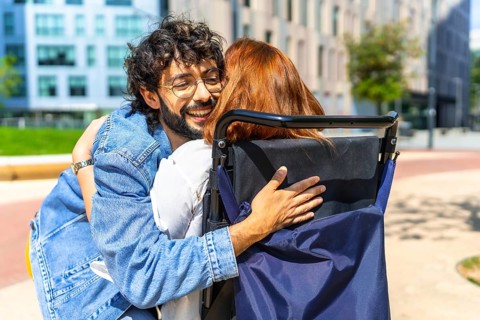 Young man with glasses is embracing a red haired woman sitting in a wheelchair on a sunny day in
