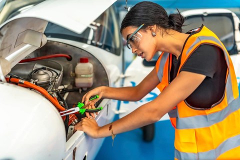 Young woman aircraft mechanic wearing safety glasses and high visibility vest using pliers while