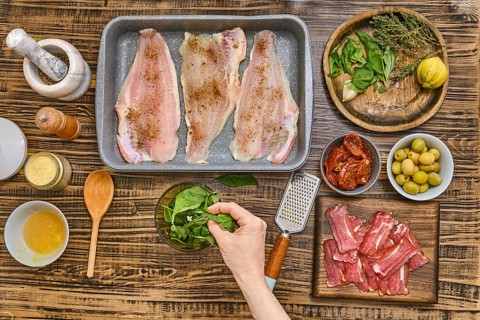 Fresh herbs, vegetables, and fish fillets are arranged on a wooden table. A hand adds spinach to a