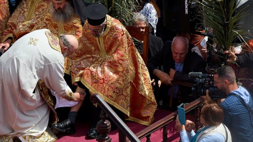 A priest in a golden robe blesses another, surrounded by cameras and a crowd, Orthodox ceremony,