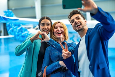 Three happy friends are taking a selfie in a hangar, making heart and victory signs, celebrating