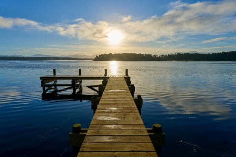 A jetty leads into a lake, the sun shines brightly and is reflected in the water, Lake Mahinapua,