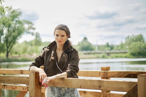 A woman stands confidently by a wooden railing in an elegant outfit, enjoying a peaceful afternoon