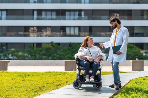 Doctor talking with smiling disabled woman in wheelchair, enjoying sunny day in urban park near