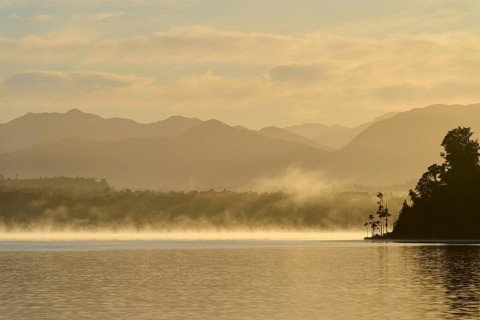A calm lake at dawn with mist, surrounded by silhouetted mountains, Lake Mahinapua, Ruatapu, South