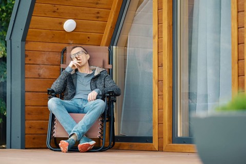 Middle-aged man sitting in lounge chair on the terrace of a modern tiny house with cihgar