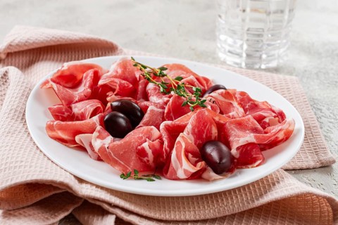 Freshly cut dried meat, served with olives and herbs, on a white plate, light background, no people