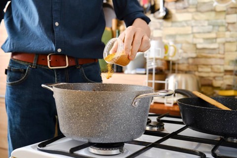 A person pouring spices into a pot on a stove. The countertop is lined with cooking utensils, and a