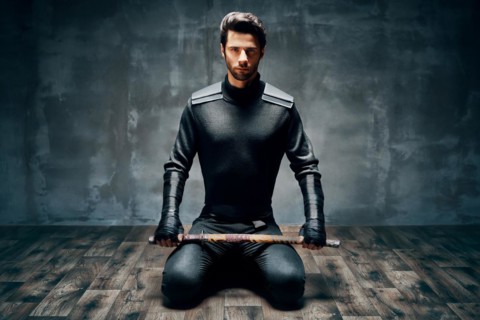 Warrior man sitting on floor posing with a sword over black background
