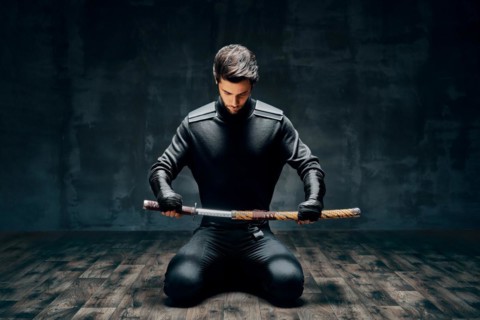 Warrior man sitting on floor posing with a sword over black background