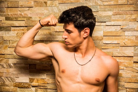Attractive and muscular shirtless young man leaning against stone wall doing bicep pose