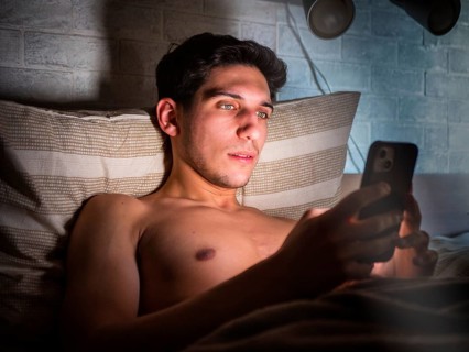 Handsome young man in bed typing on cell phone, sending text message or dialing number