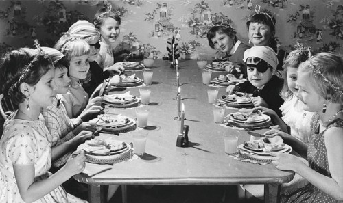 Children in the 1950s. Girls and boys dressed up in hats and costumes are sitting a table together