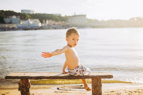 Boy child sitting on a wooden bench with his back on the beach near the water and showing his hand