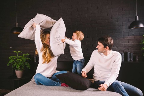 Parents with smiling child playing pillow fight on bed, happy family having fun in bedroom,