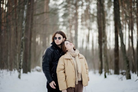 Couple in a snowy forest with the woman leaning on the man, both in winter attire among towering