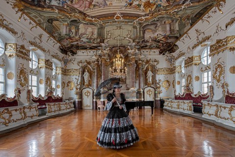 A woman in a black dress stands in a magnificent room with frescoed ceilings, staged photo shoot,