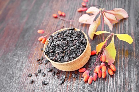 Dried black barberry in a bowl, sprigs with fresh berries and leaves on dark brown wooden board