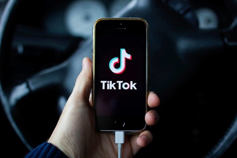 A hand holding a smartphone with TikTok app in front of a steering wheel inside a car
