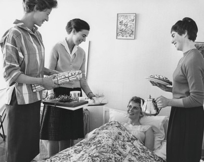 Birthday in the 1960s. A young woman lies in bed and is surprised by her girl friends singing happy