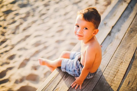 Little Caucasian boy child sitting on wooden pier sandy beach, summer time, sea holiday near water.