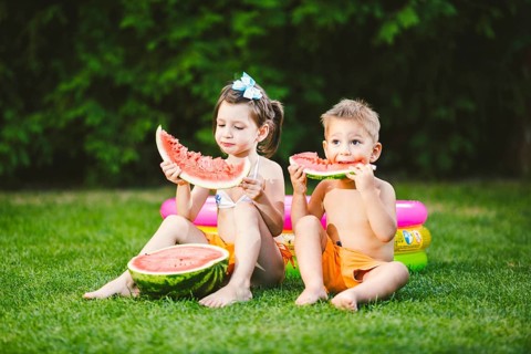 Funny little kids brother and sister eating watermelon on green graß near inflatable pool in yard