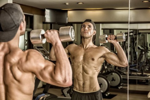 Handsome shirtless muscular young man exercising shoulders in gym with dumbbells