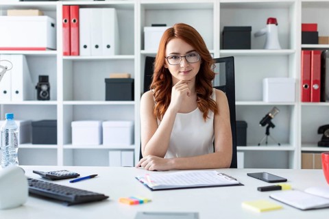 A red-haired young girl with glasses, a white blouse and a red skirt is working in the office.