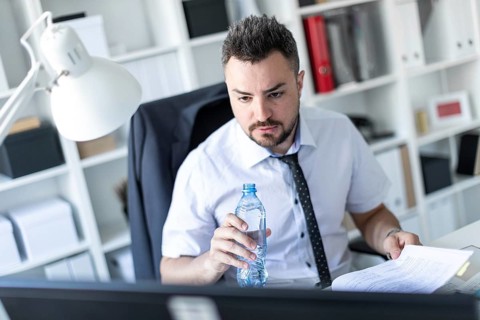 A man is sitting at a table in the office, working with documents and holding a bottle of water in