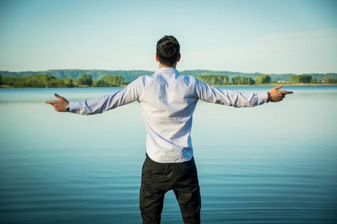 Young man in shirt, outdoor with arms spread open enjoying freedom in front of lake, seen from the