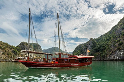 Traditional wooden boat sails on turquoise water between green hills under a cloudy sky, sailing