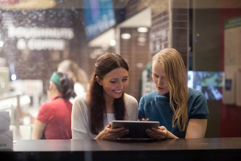 Smiling blonde and brunette women have fun in the restaurant while watching something on the tablet