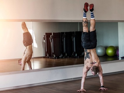 Back view of tattooed shirtless man doing handstand in sportive gym against mirror